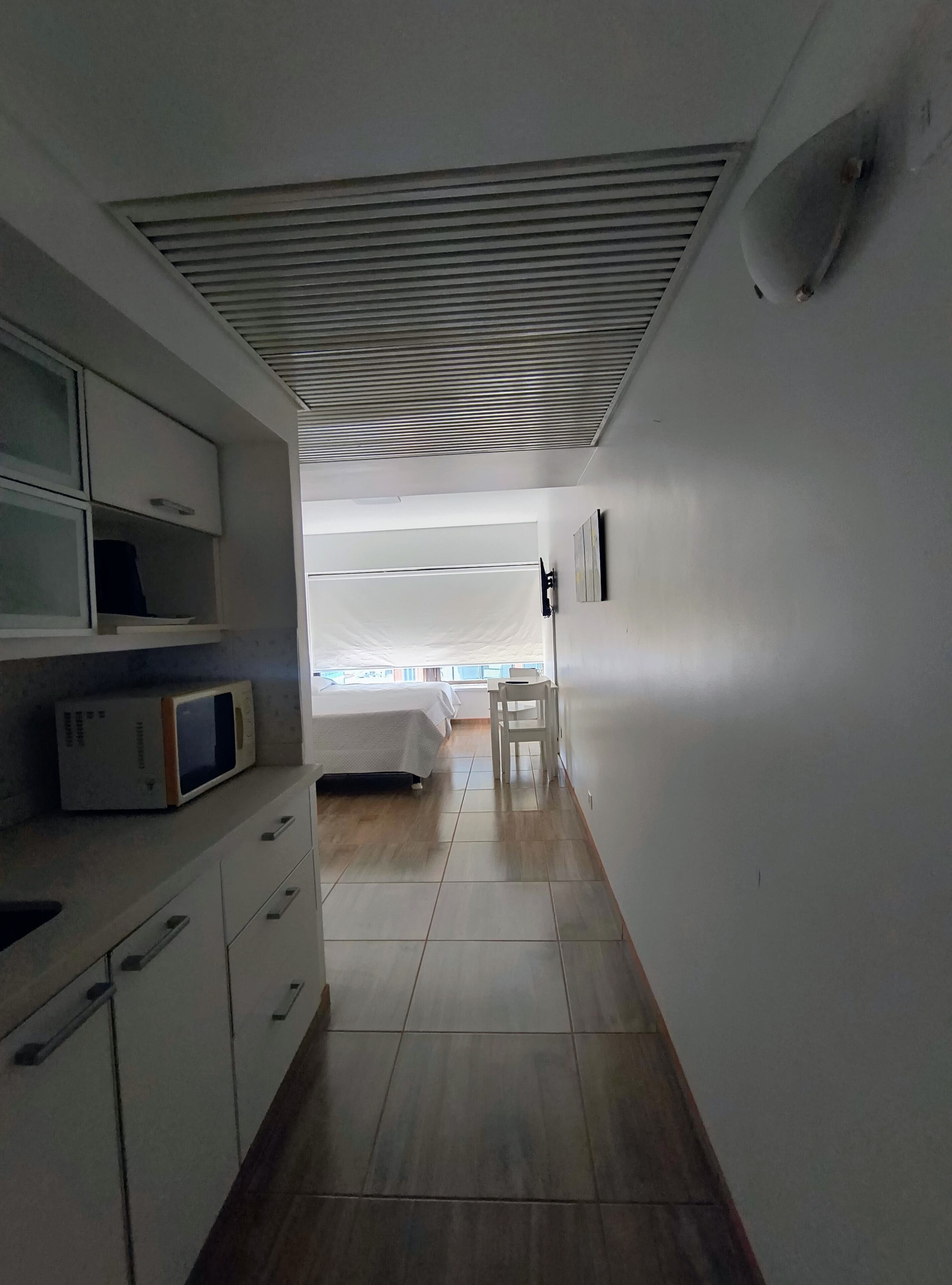 A narrow hallway leading to a bedroom with a bed, a chair, and a microwave on a cabinet.