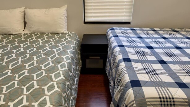 Quarto com duas camas, uma janela com persianas e uma mesinha de cabeceira.