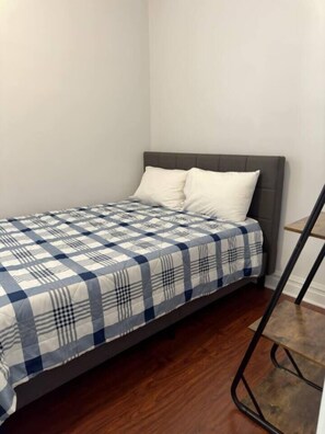 A small, single-bed room with a grey headboard, a checkered bedspread, and a wooden floor. - Spacious Apt | 4 Queen Beds | Free Parking (Chicago)