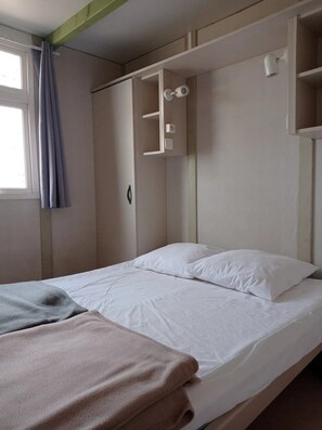 A single bed with a white mattress and pillows, a grey blanket, and a brown towel. There is a window with a curtain, a white shelf unit with a camera, and a light fixture on the wall.