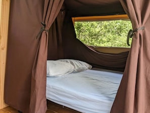 A single bed with a white mattress and pillows, set in a tent-like structure with brown curtains.