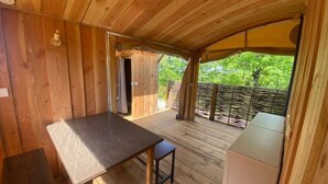 A wooden interior with a table and bench, a door leading outside, and a view of greenery through a fence.