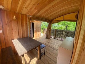 A wooden interior with a table and bench, a door leading outside, and a view of greenery through a fence.
