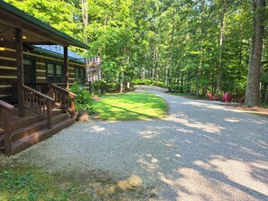 A wooden cabin with a porch, a gravel driveway, and a forested area with red chairs. - Peaceful log cabin on large acreage with fire pit and private hiking trails. (Nashville)