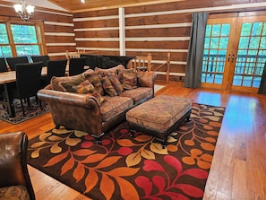 A log cabin interior with a leather sofa, a dining table with chairs, and a large window with a view of trees. - Peaceful log cabin on large acreage with fire pit and private hiking trails. (Nashville)