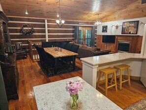 A cozy interior with a wooden dining table, a sofa, a fireplace, and a kitchen area. - Peaceful log cabin on large acreage with fire pit and private hiking trails. (Nashville)