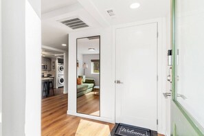 A hallway with a white door, a welcome mat, and a view into a living area with a sofa and a window. - Burien Modern Cozy - Near Airport & Amenities (Burien)