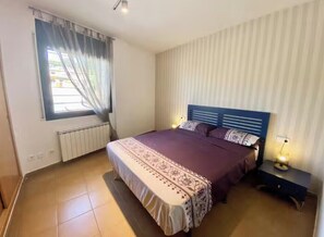 A bedroom with a bed, a nightstand, a window with curtains, and a radiator. - Apartment with a pool near the beach (Sant Feliu de Guíxols)