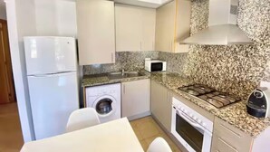 A compact kitchen with a refrigerator, microwave, washing machine, stove, and granite countertop backsplash. - Apartment with a pool near the beach (Sant Feliu de Guíxols)