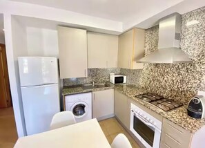 A compact kitchen with a refrigerator, microwave, washing machine, stove, and granite countertop backsplash. - Apartment with a pool near the beach (Sant Feliu de Guíxols)
