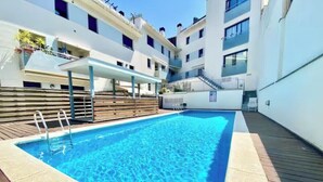 A swimming pool with a wooden deck and a covered seating area, surrounded by multi-story buildings. - Apartment with a pool near the beach (Sant Feliu de Guíxols)