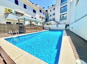 A swimming pool with a wooden deck and a covered seating area, surrounded by multi-story buildings. - Apartment with a pool near the beach (Sant Feliu de Guíxols)