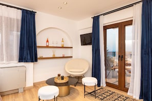 Ein modernes Wohnzimmer mit einem runden Tisch, zwei weißen Hocker, einem an der Wand montierten Fernseher und einem Balkon mit Blick.