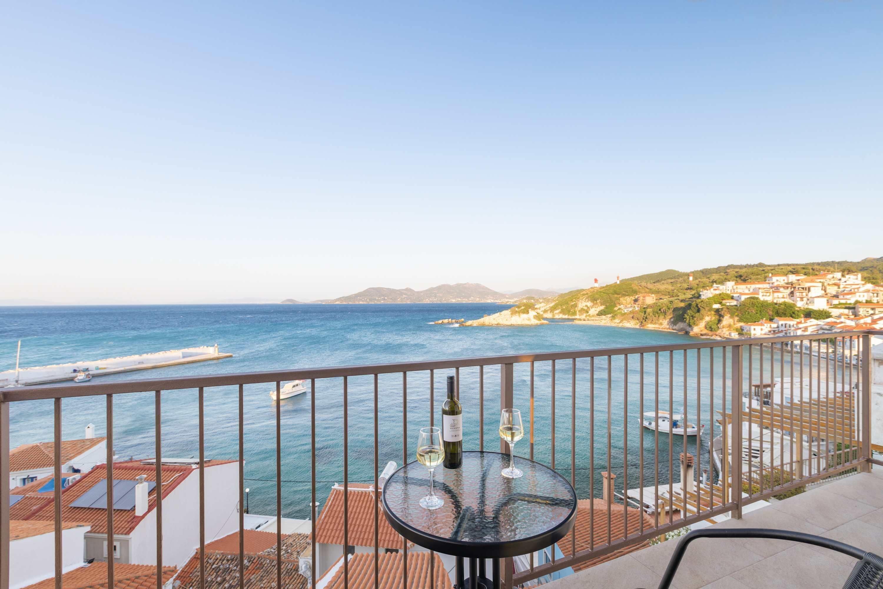 Ein Balkon mit Tisch, Flasche und Glas, mit Blick auf eine Küstenstadt, Gebäude und das Meer.