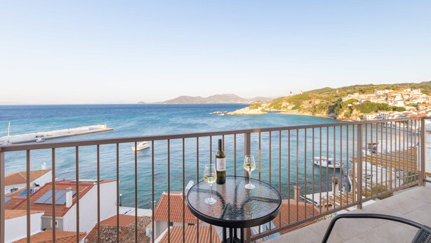 A balcony with a table, a bottle, and a glass, overlooking a coastal town with buildings and the sea. - Studio Tepes - Velis Samos Sea View Balcony Suite (Samos)