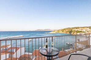 A balcony with a table, a bottle, and a glass, overlooking a coastal town with buildings and the sea. - Studio Tepes - Velis Samos Sea View Balcony Suite (Samos)