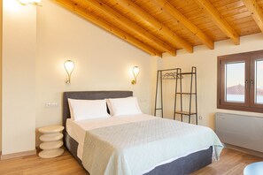 Ein Schlafzimmer mit einem Bett, Holzbalken an der Decke, Wandleuchten und einem Fenster mit Ausblick. Gegen die Wand lehnt ein Metallregal.