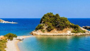 A coastal scene with a clear beach, a small boat, and a rocky outcrop with trees. - Studio Tepes - Samos Sea View Balcony Suites (Samos)