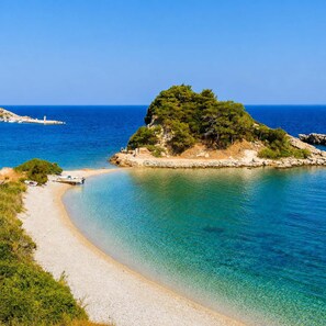 A coastal scene with a clear beach, a small boat, and a rocky outcrop with trees. - Studio Tepes - Samos Sea View Balcony Suites (Samos)