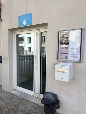 A building entrance with a reception sign, a glass door, a notice board, and a trash bin. - Appartement 3 étoiles - Piscine  - ccbgfca (Gréoux-les-Bains)