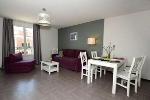 A modern living room with a sofa, a dining table with chairs, a small white table, and a hanging light fixture. - Appartement 3 étoiles - Piscine  - ccbgfcb (Gréoux-les-Bains)
