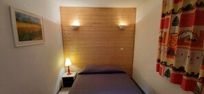 Ein kleines Zimmer mit einem hölzernen Kopfteil, einem Bett, einem Nachttisch mit Lampe und einem Bild an der Wand.