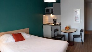 A hotel room with a bed, a small kitchenette, a round table with chairs, and a wall-mounted bulletin board. - Appartement 2 étoiles - ccbgffb (Brest)