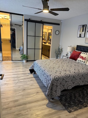 Ein Schlafzimmer mit Bett, Deckenventilator, Nachttisch und Blick zur Küche.