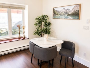 A room with a dining table, two chairs, a potted plant, a painting, and a window with a view. - Heart of North Van: 2BR Retreat with Huge Backyard (North Vancouver)