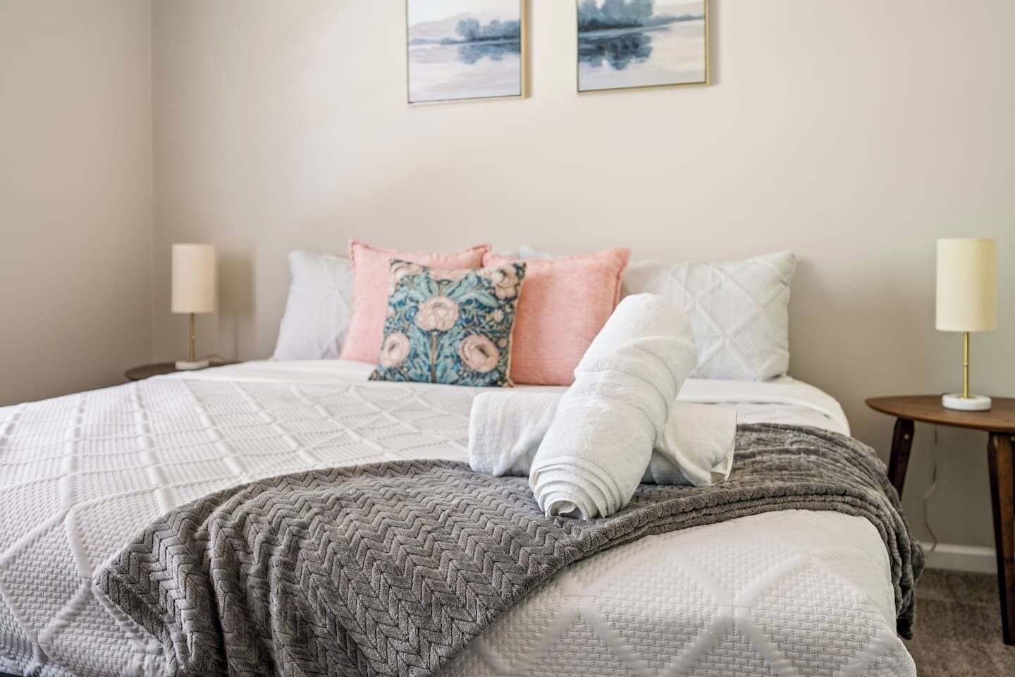 Una cama bien hecha con una manta estampada, almohadas y una toalla enrollada. Hay dos mesitas de noche con lámparas, y una obra de arte en la pared.