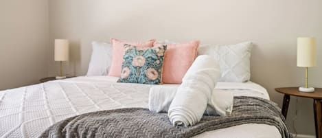 Una cama bien hecha con una manta estampada, almohadas y una toalla enrollada. Hay dos mesitas de noche con lámparas, y una obra de arte en la pared.