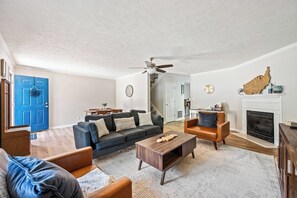 A living room with a blue door, a fireplace, a ceiling fan, and a dining area in the background.