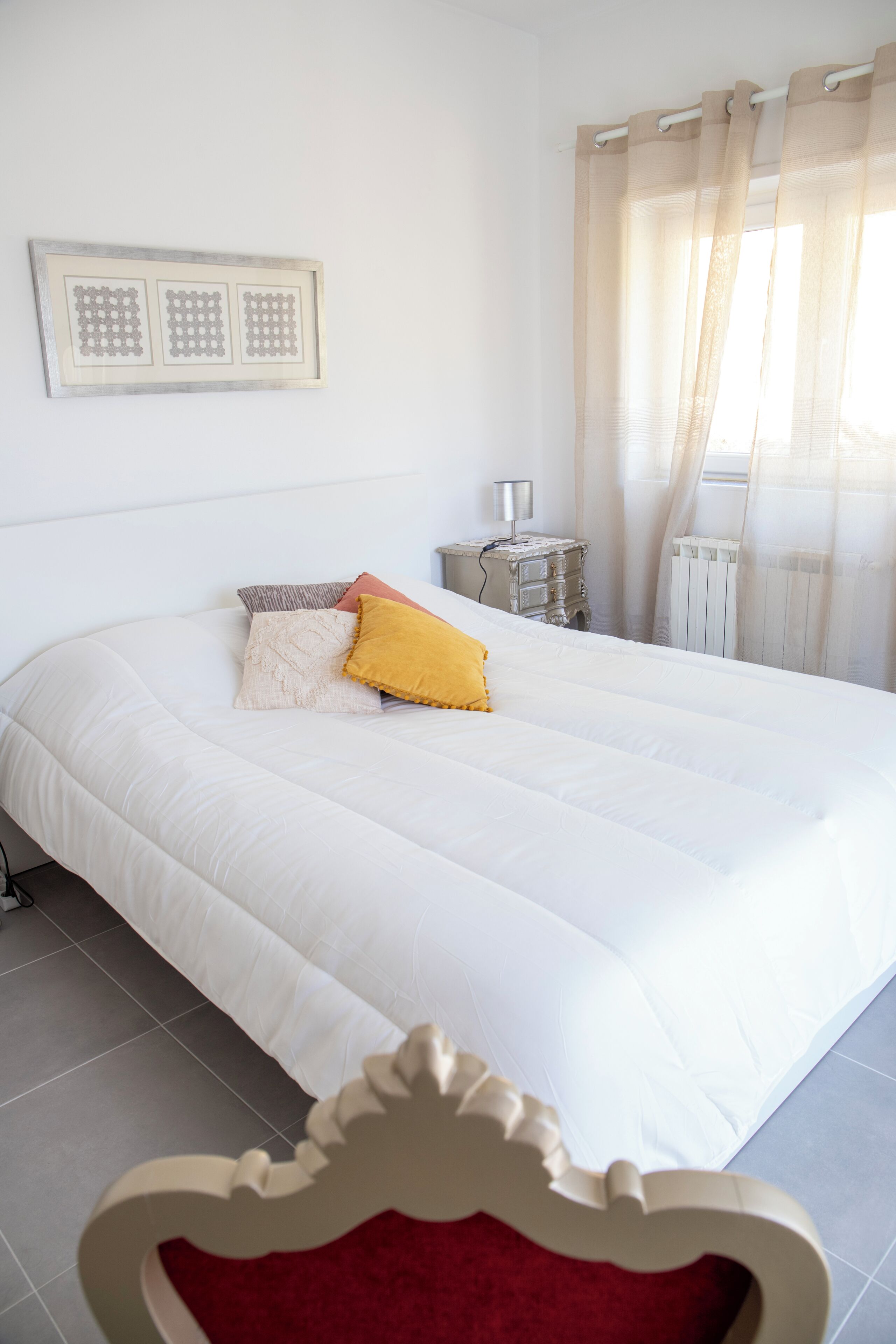 Een slaapkamer met een groot bed, een nachtkastje met een lamp, een radiator en een raam met dunne gordijnen.