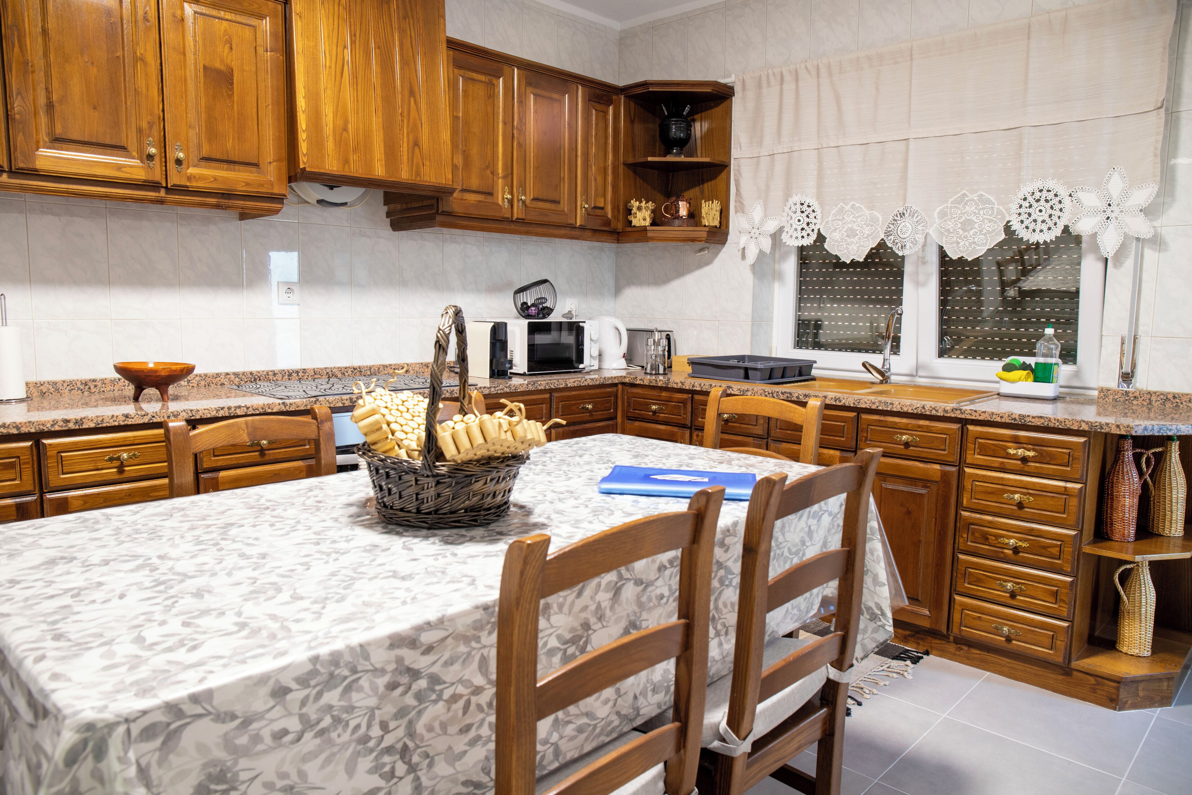 A kitchen with wooden cabinets, a marble countertop, a dining table with chairs, and a window with lace curtains.