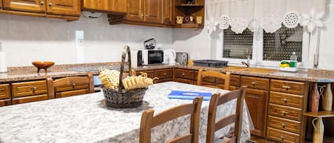 A kitchen with wooden cabinets, a marble countertop, a dining table with chairs, and a window with lace curtains.