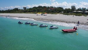Ein Strand mit angeordneten Jetskis auf dem Wasser, einem roten Luftmatratze und einem Sandstrand mit Sonnenschirmen und Menschen im Hintergrund.