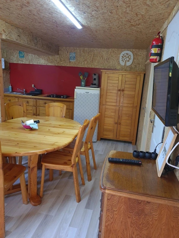 A compact kitchen with wooden cabinets, a refrigerator, a dining table with chairs, and a television on a wooden stand.