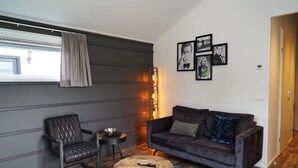 A modern living room with a leather sofa, a wooden coffee table, and a grey armchair. There are framed pictures on the wall and a window with curtains. - Cozy Kaatsheuvel Chalet (Kaatsheuvel)
