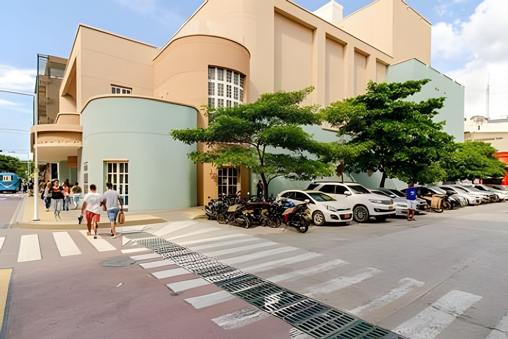 A modern building with a curved facade, a pedestrian crossing, and a parking area with multiple cars and motorcycles.