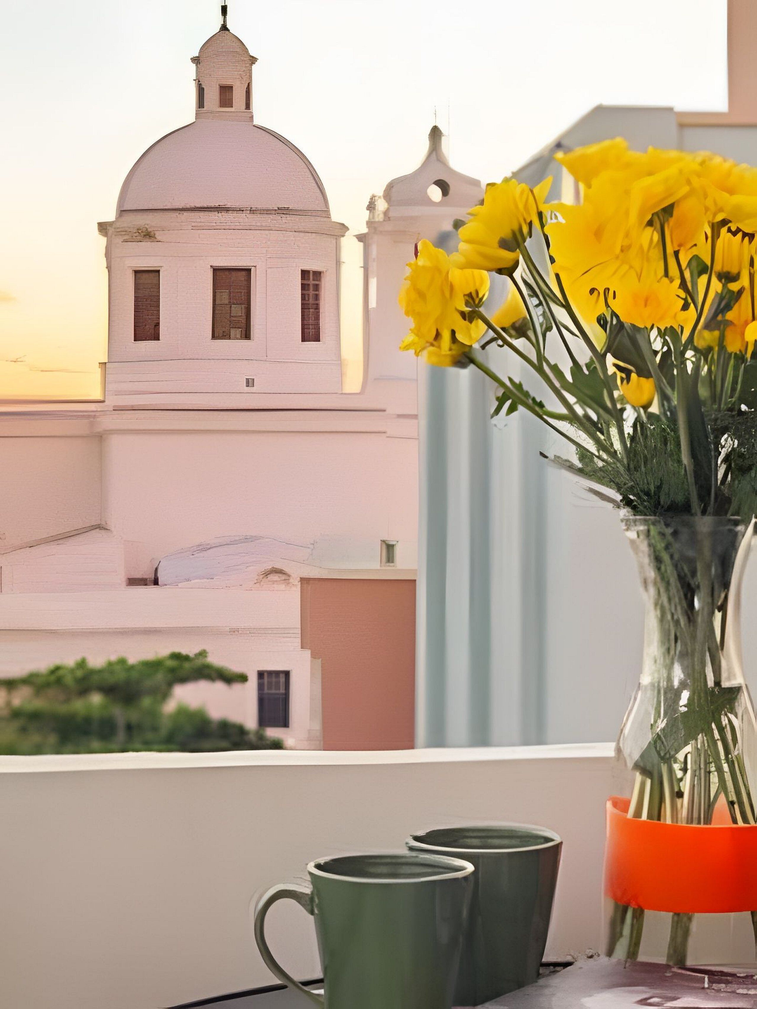 A window view of a white building with a dome and bell tower, a vase with yellow flowers, and two green mugs on a table.