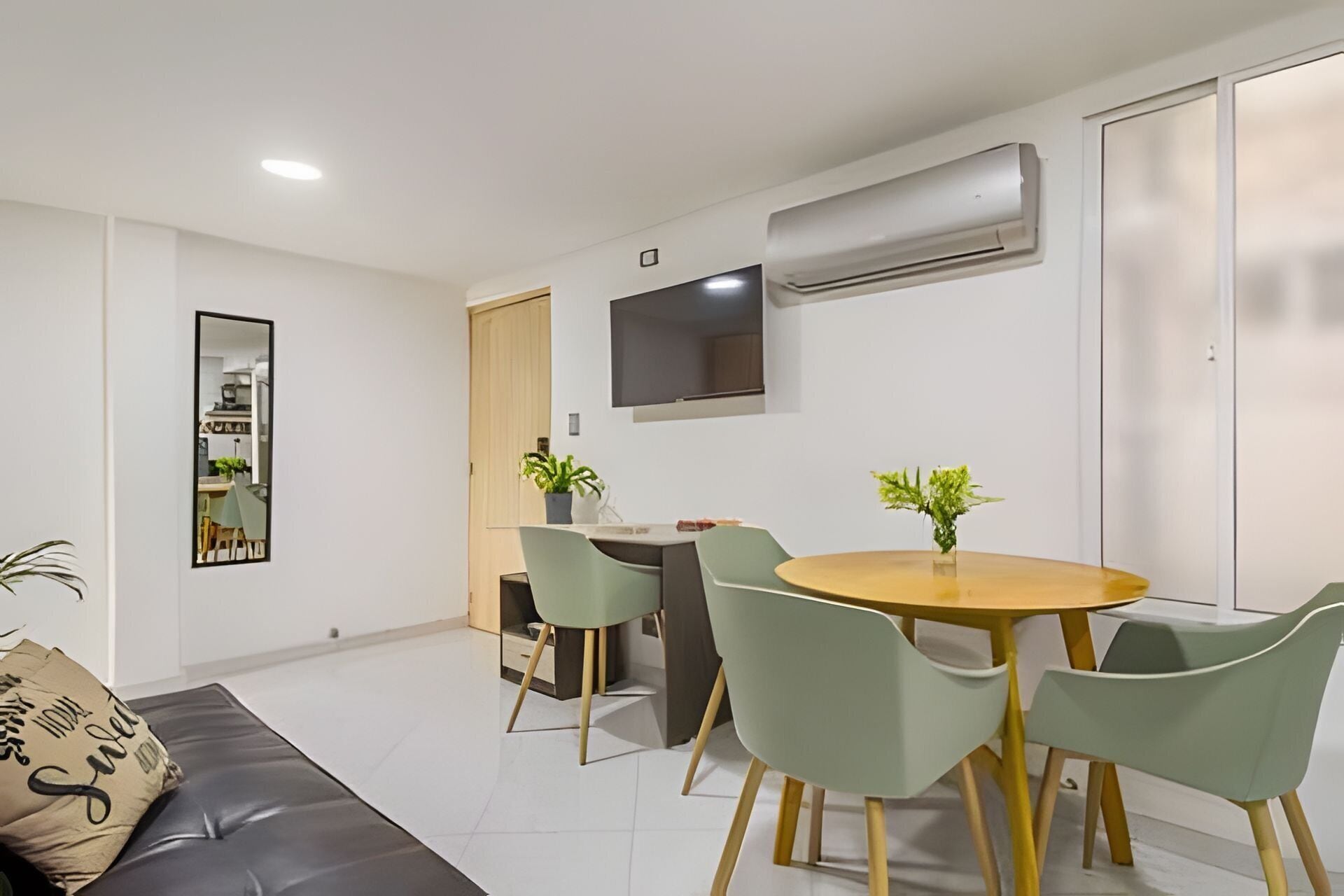 Uma sala de estar moderna com um sofá, uma mesa de jantar com cadeiras, uma TV fixada na parede e um aparelho de ar condicionado.