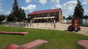 Ein Spielplatz mit Schaukeln und Rutsche, Picknicktischen und einem Gebäude mit rotem Dach.