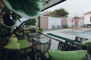 Un espacio al aire libre con mobiliario de mimbre y cojines verdes, una piscina y vistas de los edificios circundantes.