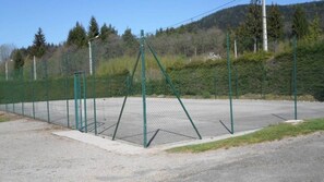 Ein durch einen Zaun abgegrenzter Tennisplatz mit Netz, umgeben von einer grünen Hecke und Bäumen im Hintergrund.