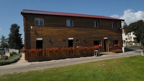 Ein zweistöckiges Holzhaus mit rotem Dach, vielen Fenstern und einer kleinen Außenbestuhlung mit roten Blumen.