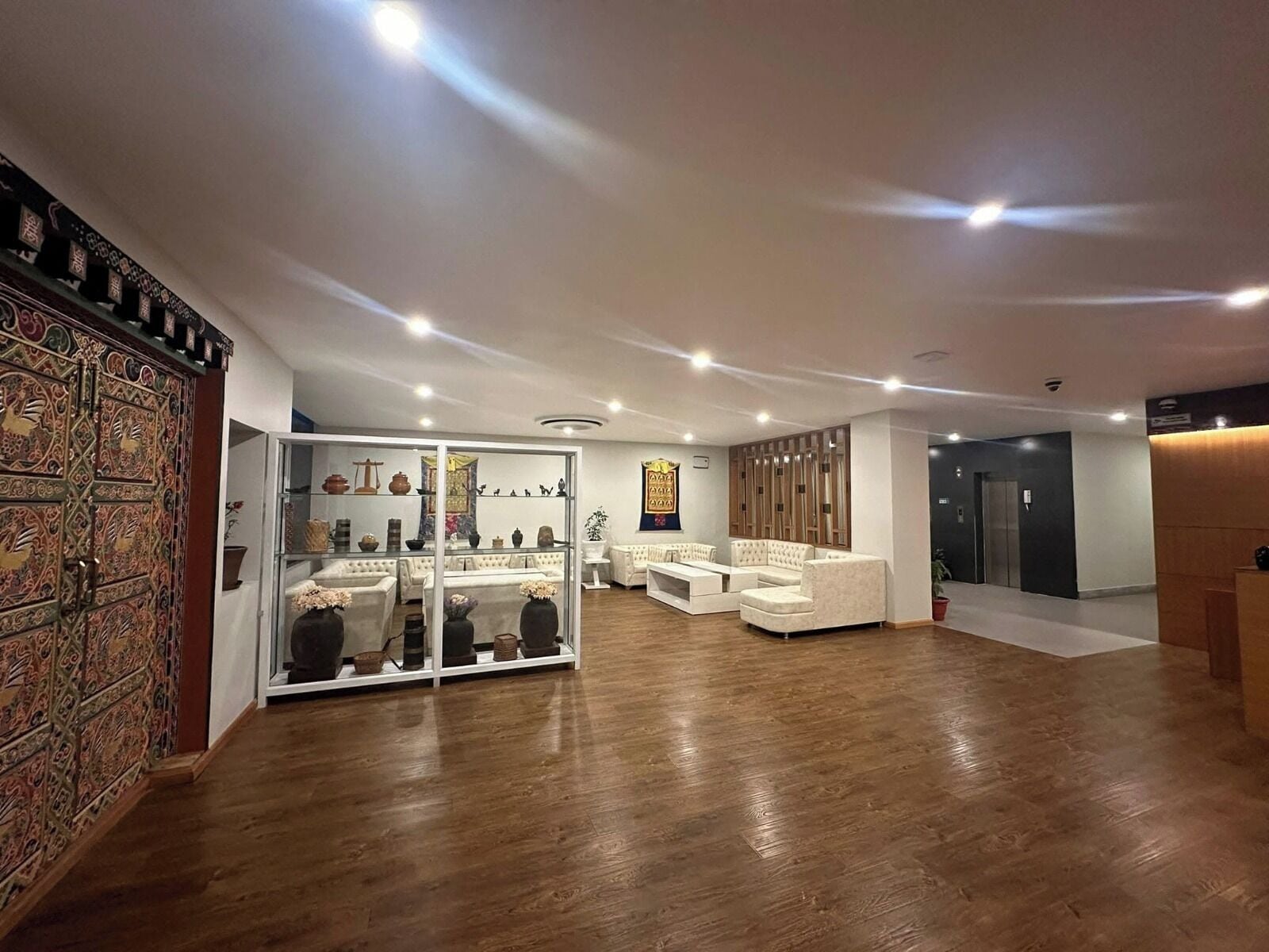 Um hall amplo com design moderno, apresentando um painel de vidro com objetos decorativos, área de estar com sofás e painel de parede em madeira.