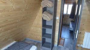 Quarto com paredes de madeira, uma cama com colchão cinza, uma prateleira com toalhas dobradas e uma porta que dá para outro quarto.