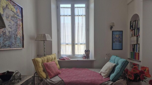 Un dormitorio con cama, ventana con cortinas, mesita de noche, lámpara, silla, estantería y un cuadro.