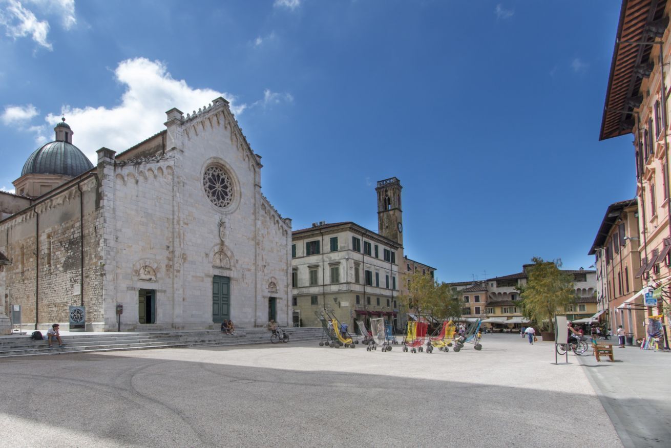 A historic building with a dome and bell tower, surrounded by a plaza with flags and a few people.