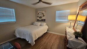Ein Schlafzimmer mit Bett, Deckenventilator, Nachttisch mit Lampe und einem Fenster mit Jalousien.